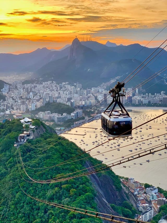 Una funivia percorre una montagna verde e rigogliosa, dominando una città costiera, una baia piena di barche e le montagne al tramonto.
