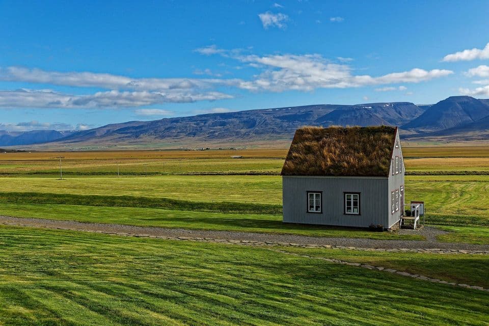 Una casa grigia con un tradizionale tetto in torba si erge solitaria in un vasto campo verde, con montagne distanti sotto un cielo blu.