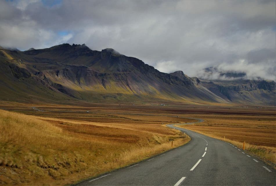 Una strada asfaltata sinuosa si snoda attraverso un vasto paesaggio dorato, dirigendosi verso un'imponente catena montuosa sotto un cielo nuvoloso.