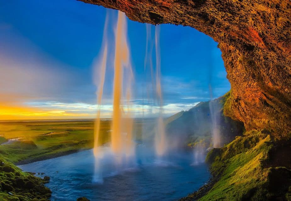Vista da dietro una cascata impetuosa che si affaccia su un paesaggio verde e un fiume durante un tramonto vivace.