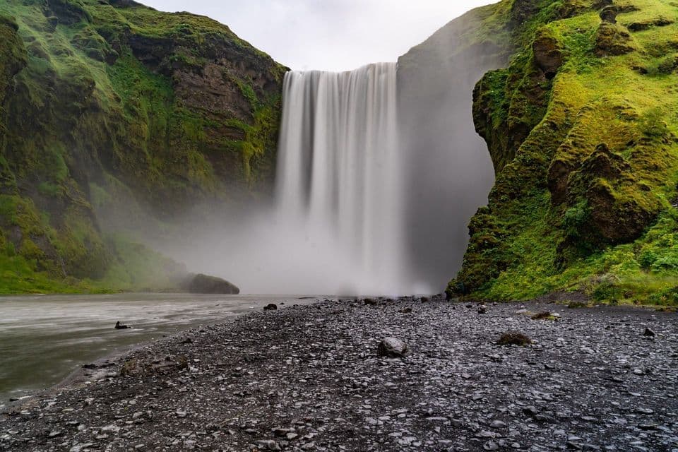 Uno scatto a lunga esposizione di una vasta cascata che si getta tra scogliere verdi e muschiose su una riva scura e rocciosa.
