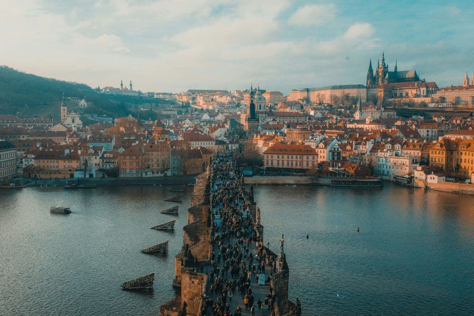 Una vista dall'alto di un ponte di pietra affollato su un fiume, con un panorama urbano europeo e un castello sullo sfondo.