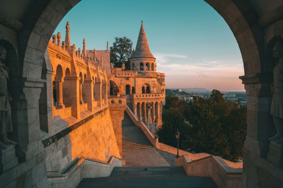 Una vista da un arco in pietra di un bastione storico con torrette, illuminato dalla luce dorata dell'alba su un panorama urbano.