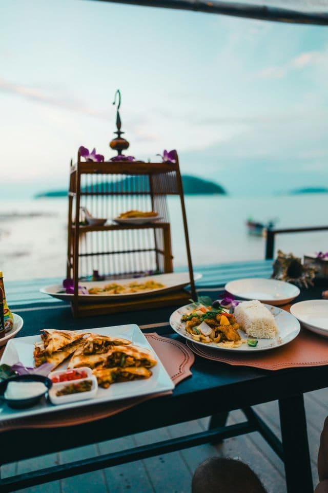 Piatti di cibo disposti su un tavolo con un supporto da portata decorativo in legno, con vista sull'oceano al tramonto.
