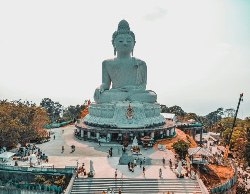 Vista aerea di una massiccia statua bianca del Buddha seduto su una collina, con persone che visitano la piazza e i giardini sottostanti.
