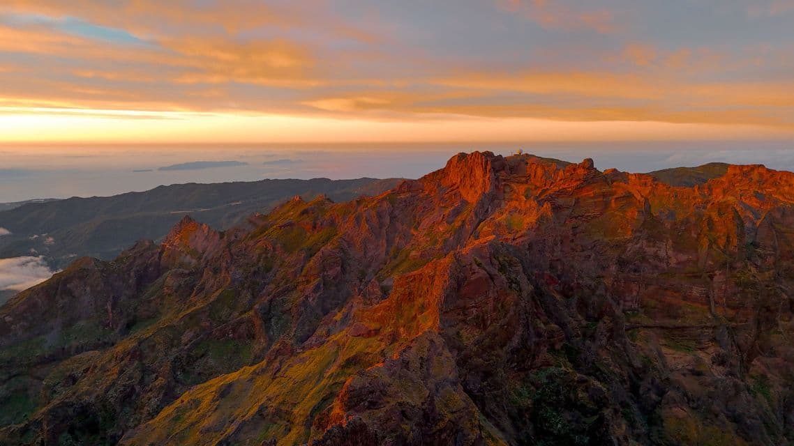 Una vista aerea di aspre vette montuose che risplendono nella luce dorata dell'alba, con un cielo nuvoloso sull'oceano.