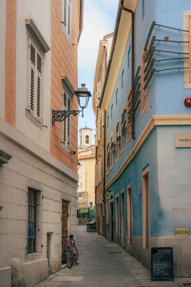 Un vicolo stretto acciottolato tra edifici colorati, con una bicicletta rossa parcheggiata e un campanile visibile in lontananza.