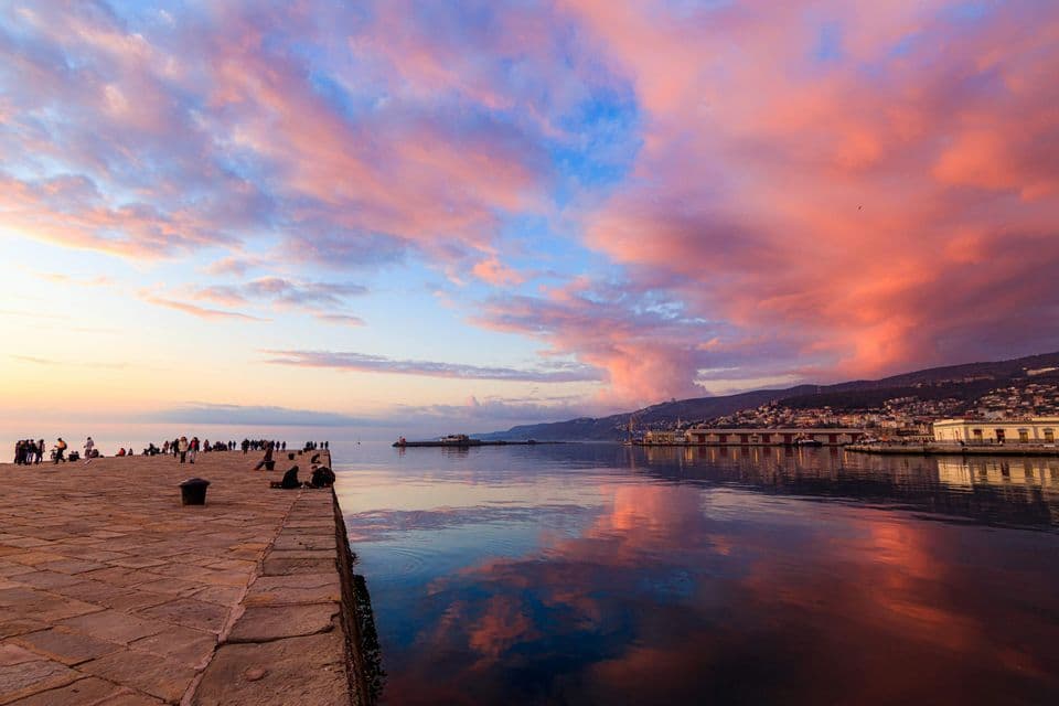Le persone si riuniscono su un molo di pietra che si affaccia su un porto calmo al tramonto, con nuvole rosa che si riflettono nell'acqua.