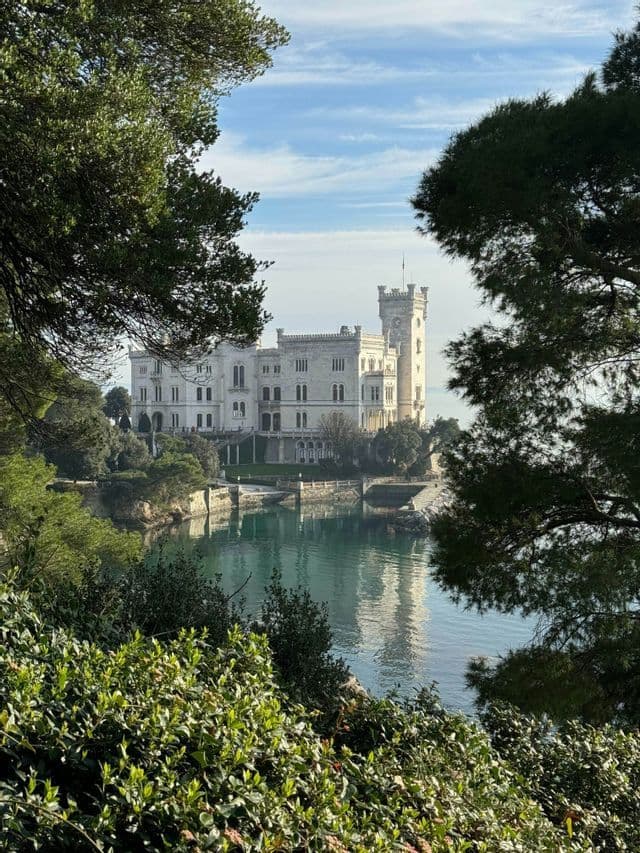 Un castello bianco con una torre si staglia sulla riva di acque calme, incorniciato da alberi e arbusti verdi.