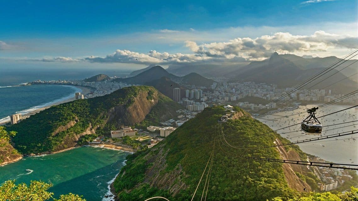 Una vista aerea da una funivia che domina una città costiera, una spiaggia curva e montagne verdi lussureggianti sotto un cielo parzialmente nuvoloso.