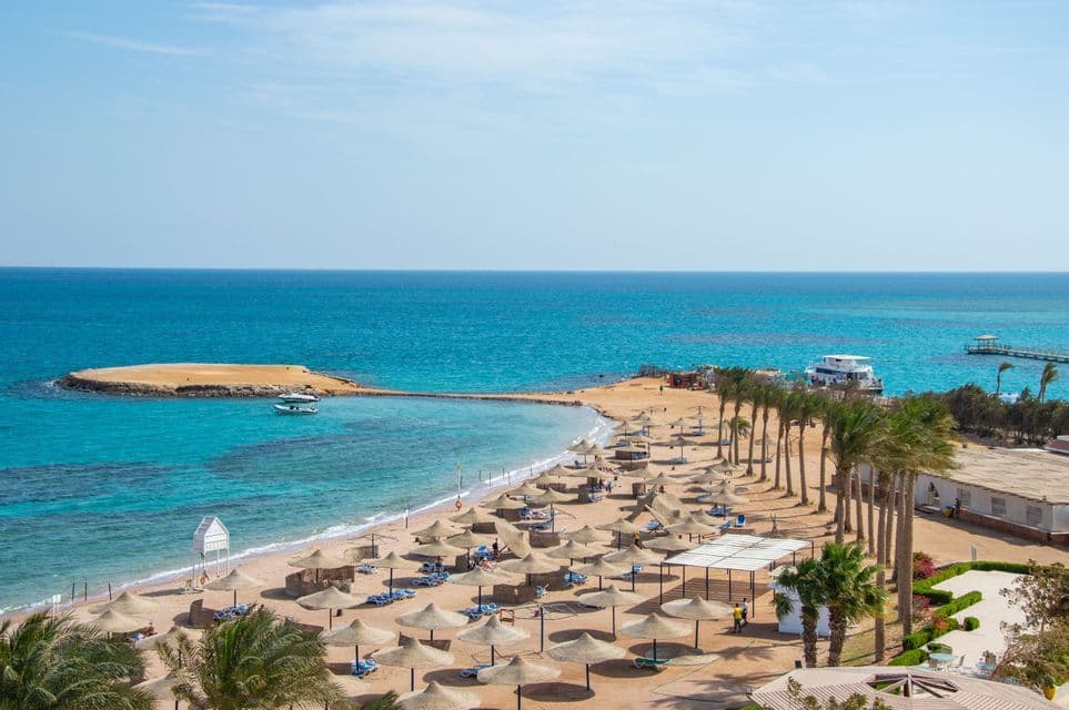 Una vista sopraelevata di un resort balneare sabbioso con molti ombrelloni di paglia e palme accanto ad acque turchesi.