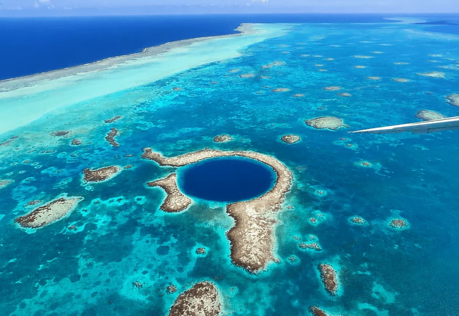 Vista aerea di una grande voragine blu scuro, circondata da un atollo corallino in un mare turchese poco profondo, con un'ala di aereo visibile.