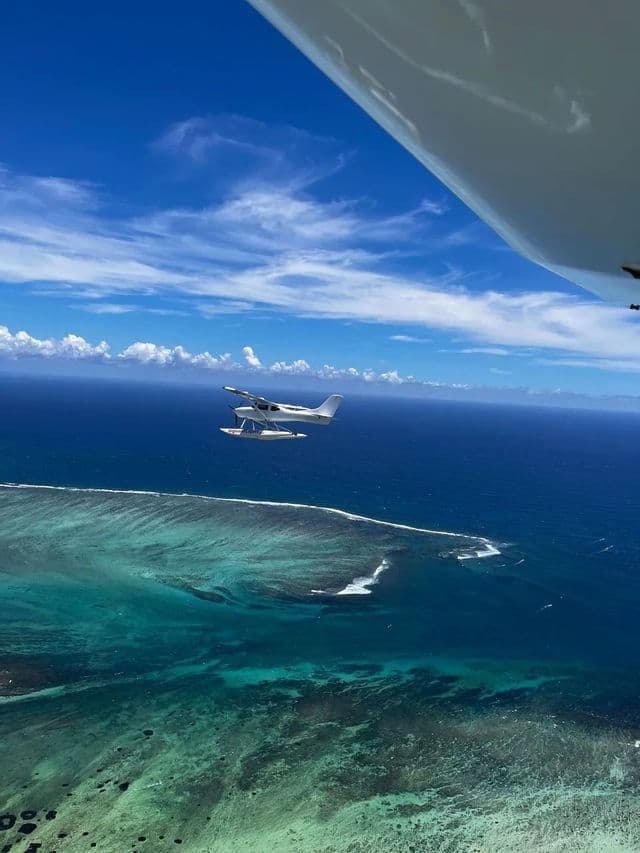 Un idrovolante bianco vola sopra una barriera corallina turchese e un oceano blu profondo sotto un cielo parzialmente nuvoloso, visto dall'ala di un altro aereo.