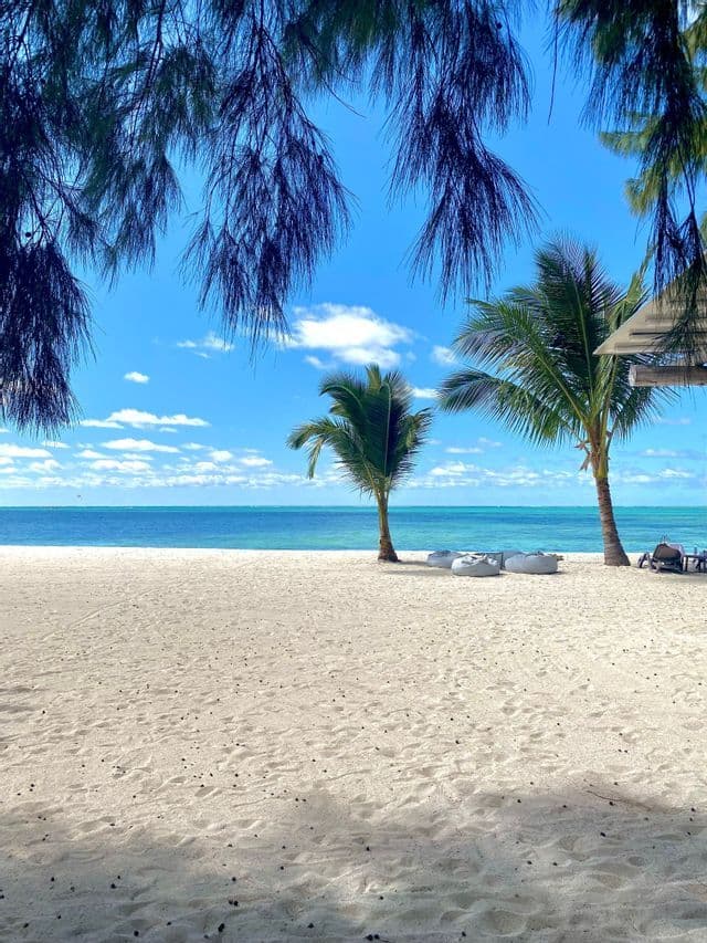 Palme e pouff su una spiaggia di sabbia bianca, affacciata su un oceano turchese, sotto un cielo azzurro e parzialmente nuvoloso.
