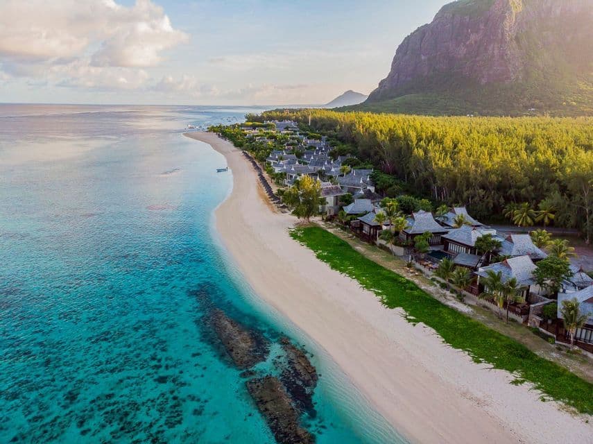 Una vista aerea di un resort su una spiaggia di sabbia bianca accanto ad acque turchesi, con una foresta lussureggiante e un'imponente montagna dietro.