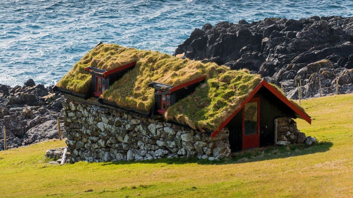 Una casa tradizionale in pietra con un tetto erboso si trova su una collina verde accanto a una costa rocciosa e al mare.