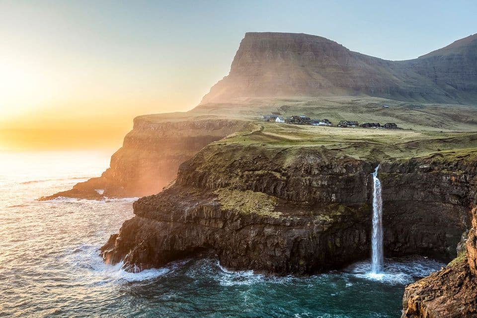 Una cascata si getta da una scogliera verde direttamente nell'oceano, con un piccolo villaggio visibile sullo sfondo al tramonto.