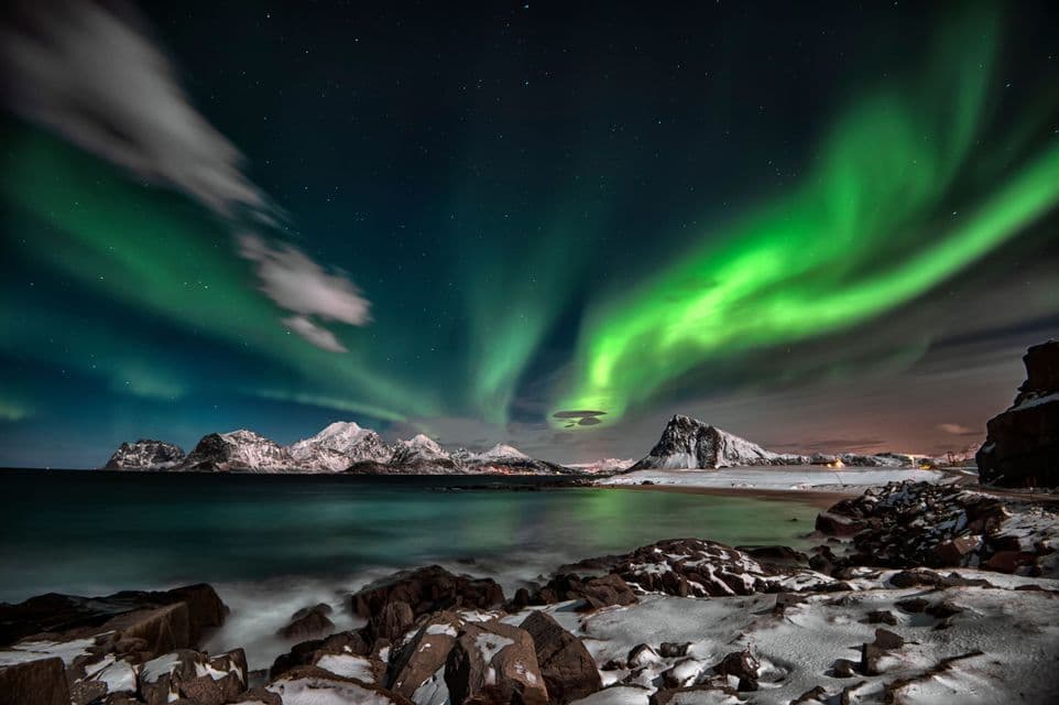 L'aurora boreale verde danza nel cielo notturno stellato sopra montagne innevate e una costa rocciosa e innevata.