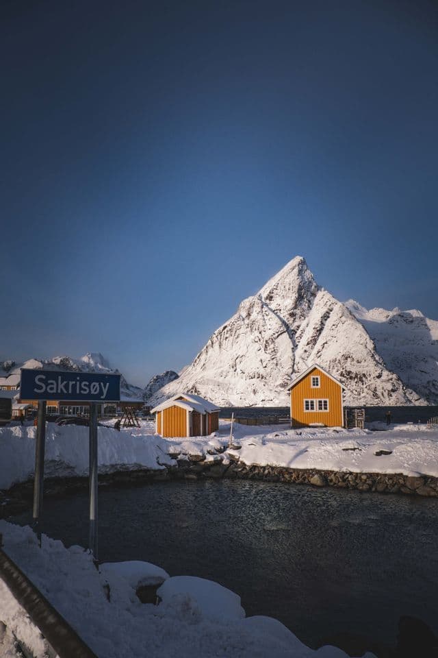 Capanne gialle sorgono su una riva innevata di fronte a un'imponente montagna frastagliata ricoperta di neve, sotto un cielo azzurro e limpido.