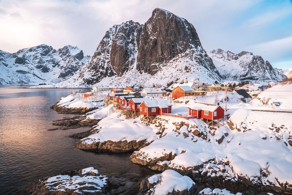 Un villaggio di case rosse si trova su una costa innevata ai piedi di grandi e ripide montagne vicino all'acqua.