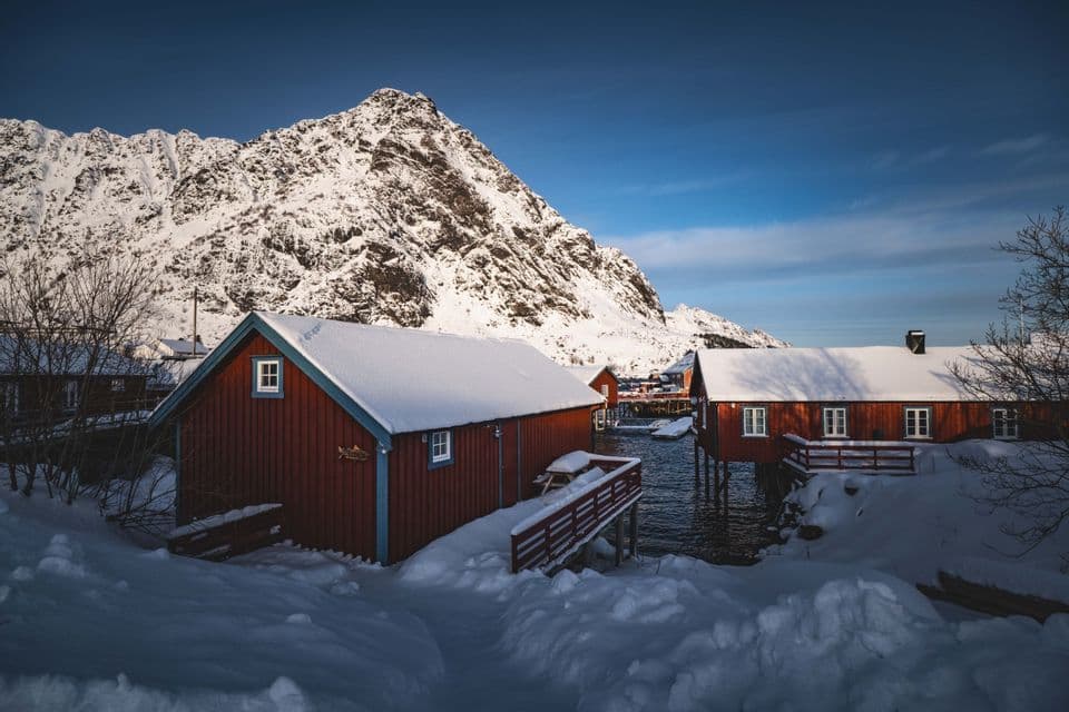 Case di legno rosse con tetti innevati si trovano in riva all'acqua, ai piedi di una grande montagna innevata, sotto un cielo azzurro e limpido.