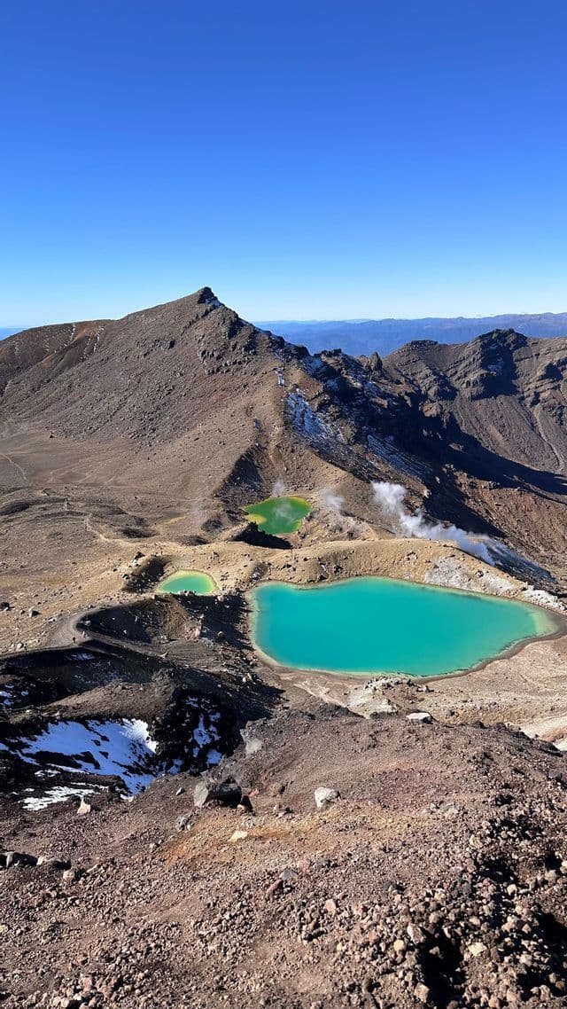 Una veduta dall'alto di tre laghi di cratere colorati, uno turchese e due verdi, in una catena montuosa rocciosa e vulcanica sotto un cielo azzurro e limpido.