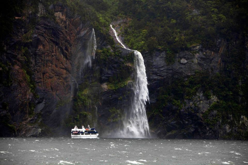 Un battello turistico naviga su acque agitate ai piedi di un'alta cascata che precipita da una ripida parete rocciosa ricoperta di vegetazione verde.