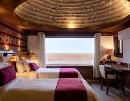 Une chambre d'hôtel avec deux lits simples et un plafond voûté et texturé donne sur un paysage désertique par une grande fenêtre.