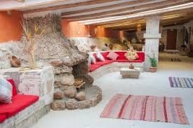 Un salon rustique avec une grande cheminée en pierre, un canapé rouge incurvé et un plafond avec poutres apparentes.