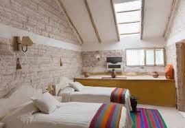 Une chambre d'hôtel rustique avec deux lits simples, des murs en briques blanches texturées et un plafond mansardé avec une lucarne.