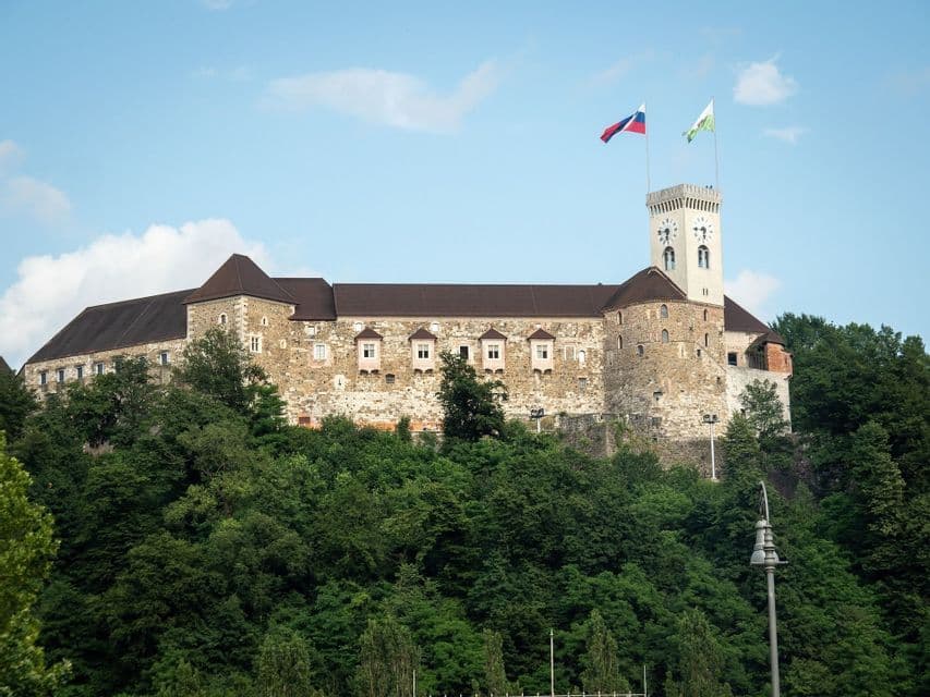 Un castillo de piedra histórico con una torre de reloj se alza sobre una colina densamente arbolada bajo un cielo azul con nubes ligeras.