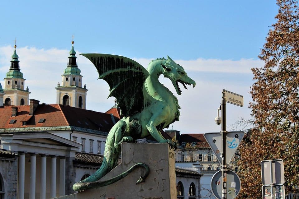 Una estatua de un dragón verde con alas abiertas se alza sobre un pedestal de piedra, con edificios históricos y campanarios de iglesias al fondo.