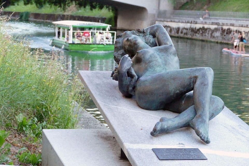 Una escultura de bronce de una mujer reclinada sobre un pedestal de piedra junto a un río, con un barco turístico y practicantes de paddleboard al fondo.