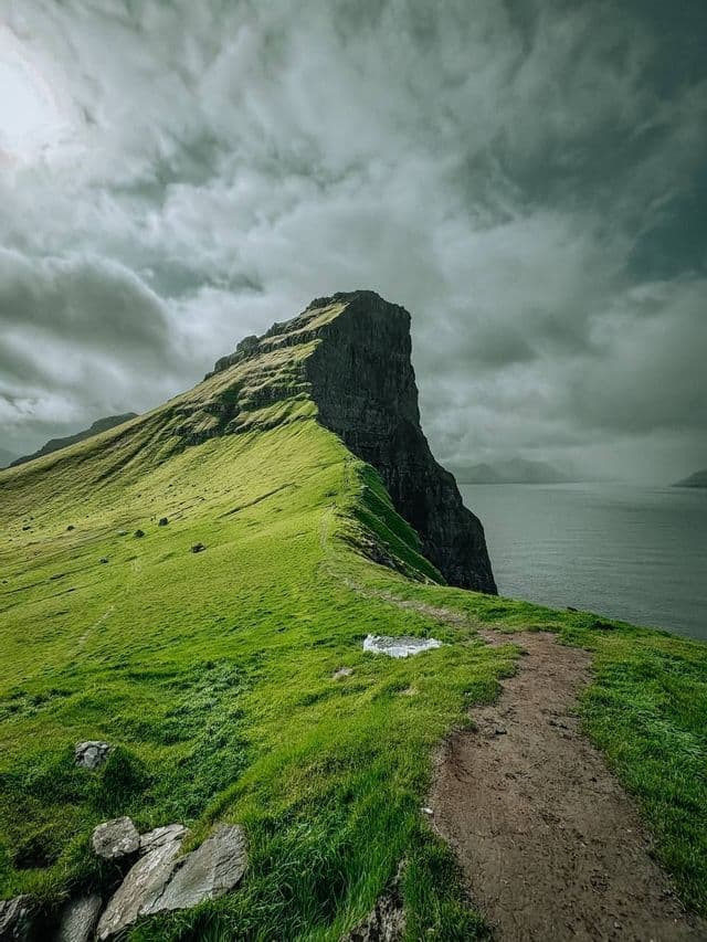 Un sentiero sterrato si snoda lungo un ripido crinale montuoso verde che si affaccia sul mare sotto un cielo nuvoloso.