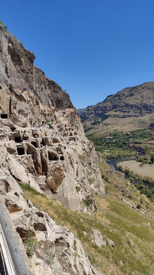 Un complesso di grotte scavate in una ripida scogliera si affaccia su una verde valle fluviale sotto un cielo azzurro e limpido.