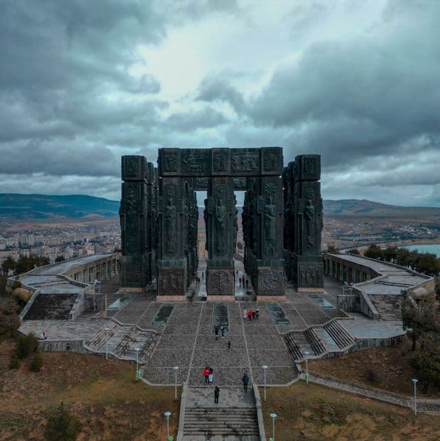 Una veduta aerea di un imponente monumento scolpito su una collina, con persone che camminano nella piazza sottostante sotto un cielo nuvoloso.