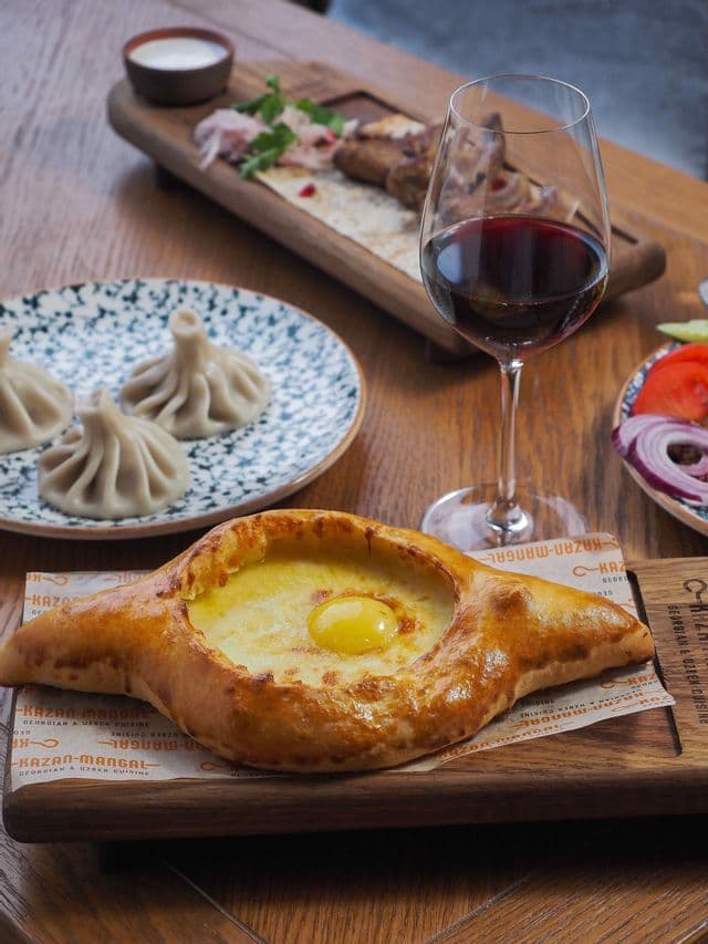 Un pasto tradizionale georgiano con khachapuri, khinkali e un bicchiere di vino rosso disposti su un tavolo di legno.