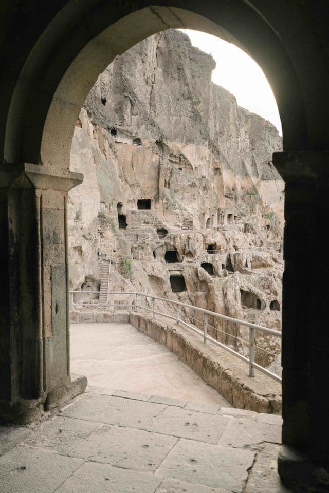 Vista da un arco di pietra di una passerella che conduce a un grande complesso di grotte scavate in una scogliera rocciosa a picco.