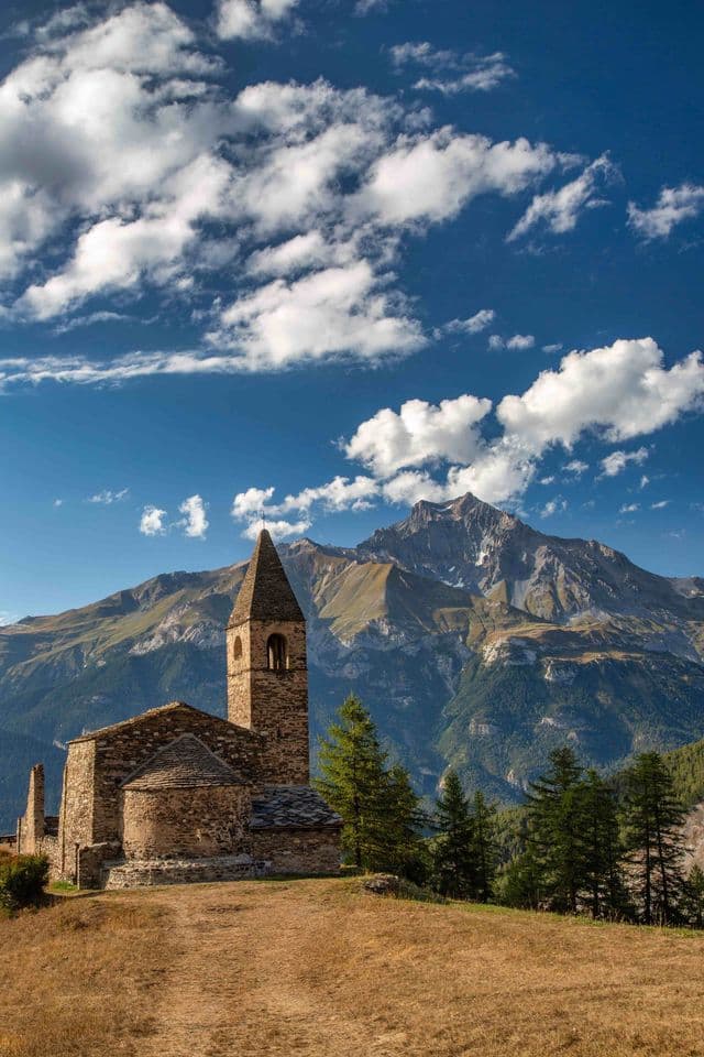 Una chiesa rustica in pietra con campanile si erge su una collina erbosa, con un'ampia catena montuosa sullo sfondo sotto un cielo blu e nuvoloso.