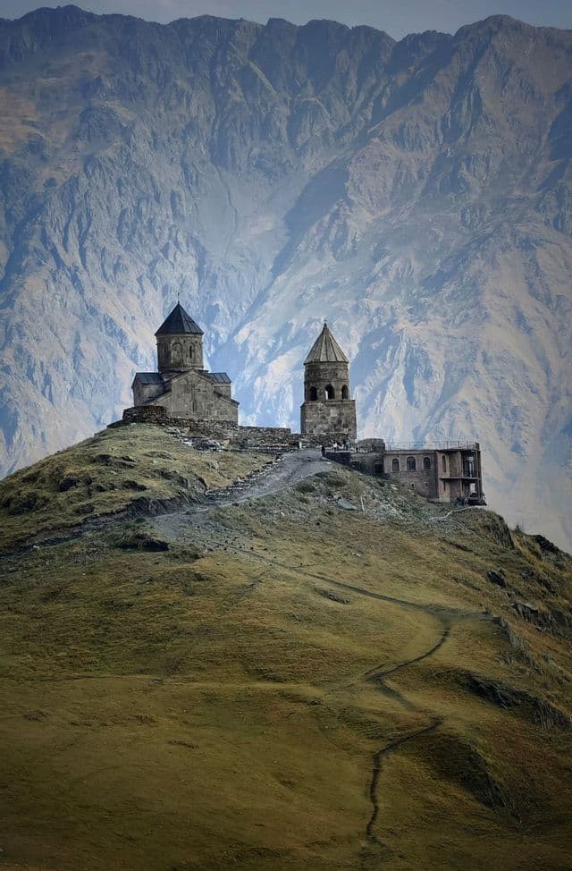 Una chiesa in pietra e un campanile si ergono su una collina verdeggiante, con una maestosa catena montuosa che si staglia sullo sfondo.
