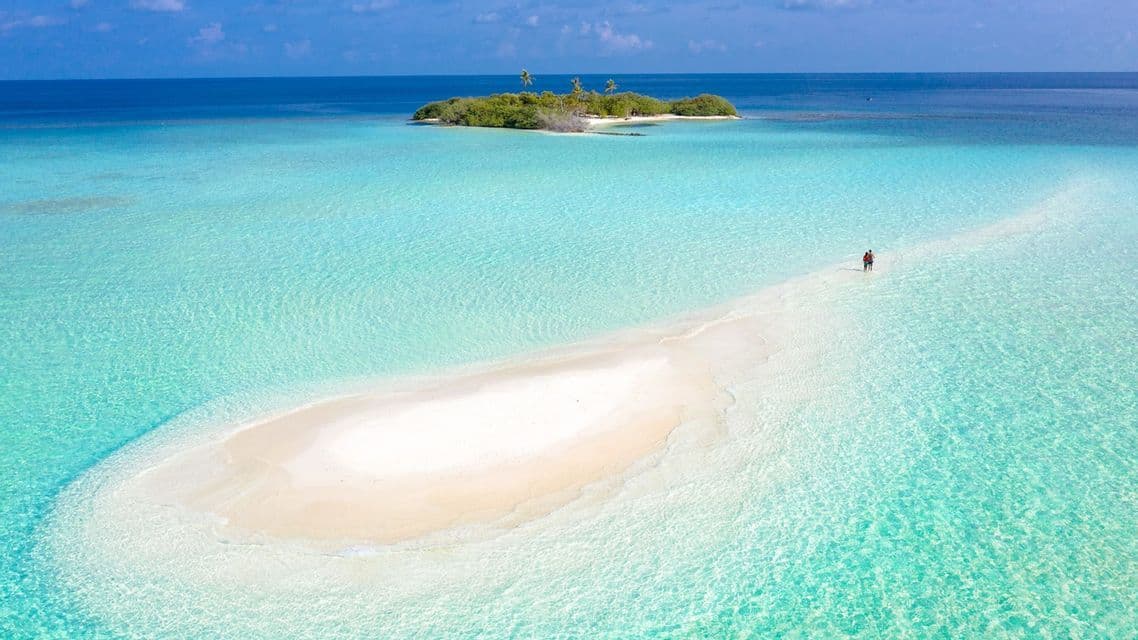 Veduta aerea di due persone su un banco di sabbia bianca che serpeggia in acque turchesi verso una piccola isola tropicale.