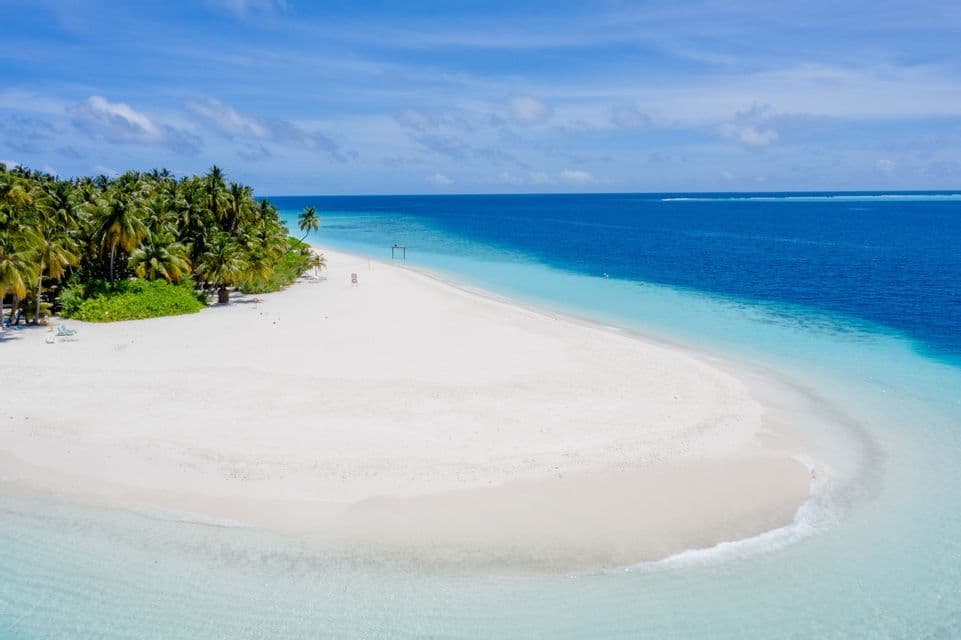 Una veduta aerea di una lingua di sabbia bianca che si estende da un'isola tropicale con palme nell'oceano turchese e blu.