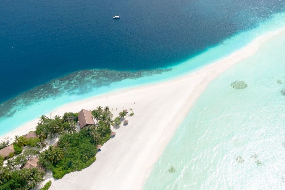 Una vista aerea di una striscia di sabbia bianca con palme e capanne, che separa l'oceano blu profondo dall'acqua turchese bassa.