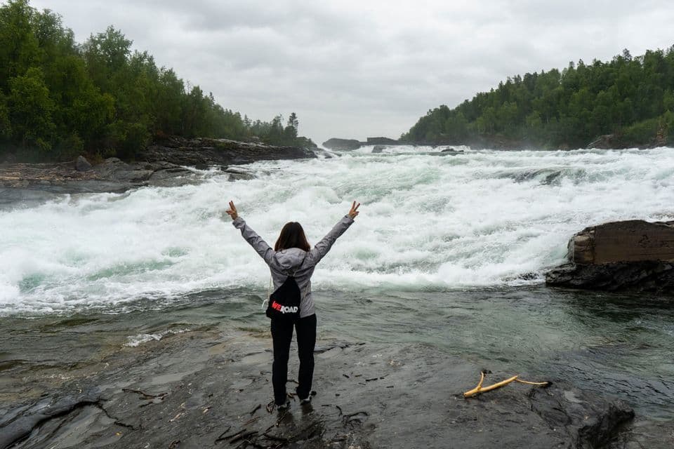 Una persona en un viaje de WeRoad se encuentra en una orilla rocosa con los brazos levantados, frente a un río turbulento con rápidos de aguas bravas.
