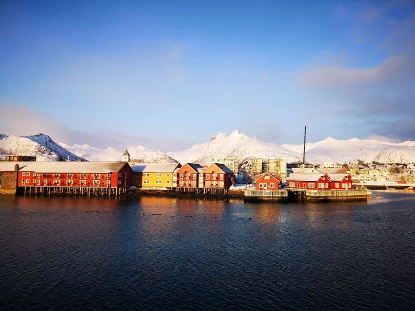 Bunte rot-gelbe Stelzenhäuser säumen einen ruhigen Hafen mit großen, schneebedeckten Bergen im Hintergrund unter blauem Himmel.