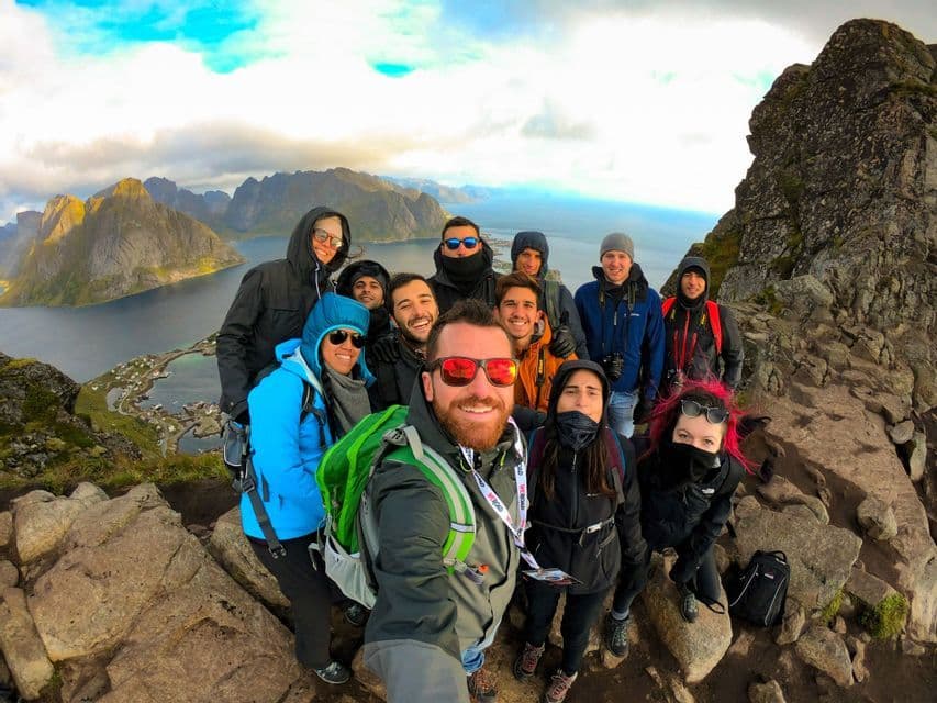 Un groupe WeRoad en voyage prend un selfie au sommet d'une montagne rocheuse, avec vue sur un village côtier et des montagnes de l'autre côté d'une baie.