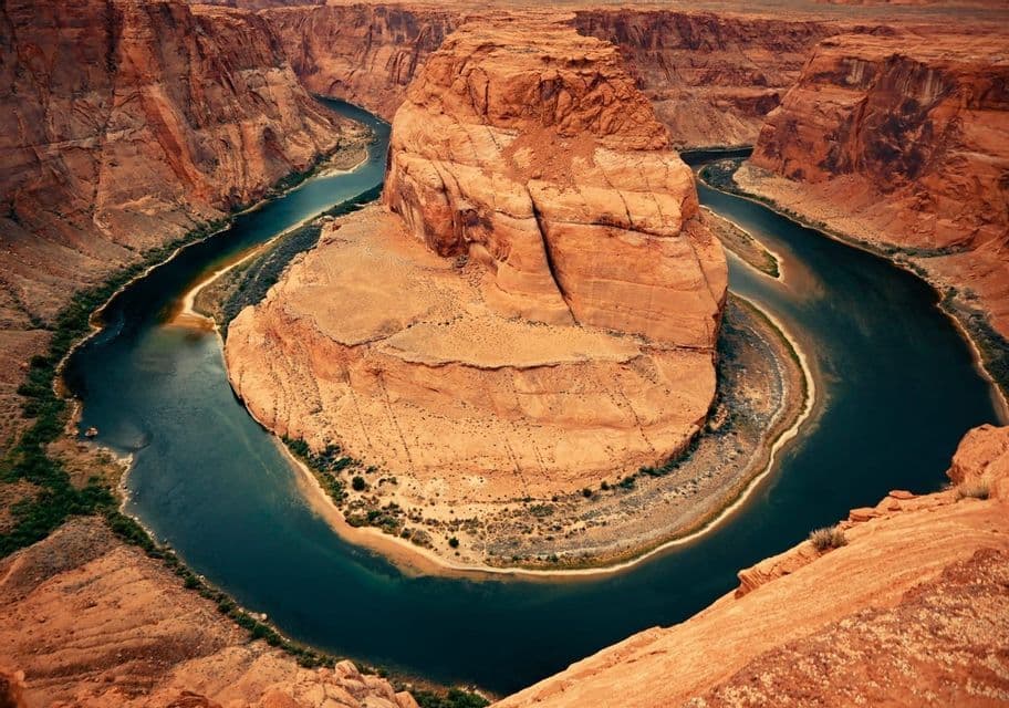 Un fiume blu scuro scorre in una curva a ferro di cavallo attorno a una grande formazione rocciosa centrale, all'interno di un profondo canyon di roccia rossa.