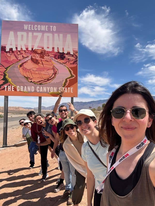 Un gruppo WeRoad in viaggio posa per un selfie in fila accanto al cartello 'Welcome to Arizona'.