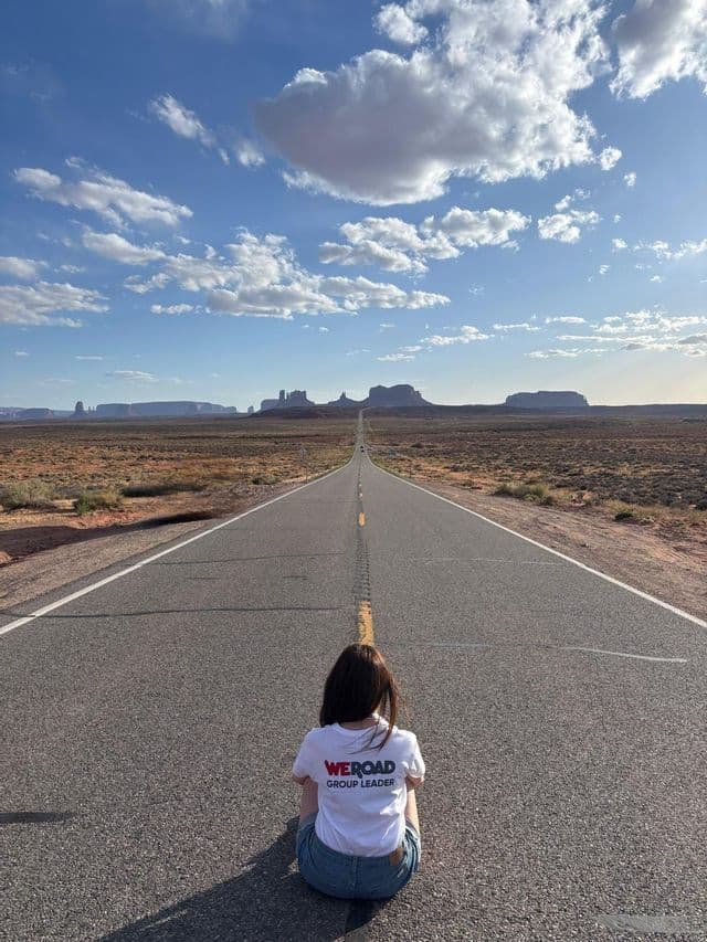 Un capogruppo WeRoad è seduto nel mezzo di una lunga strada nel deserto, che guarda verso formazioni rocciose all'orizzonte.