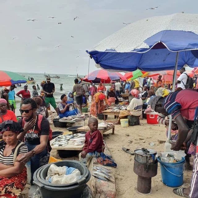 Un vivace mercato del pesce su una spiaggia sabbiosa, con molte persone riunite sotto ombrelloni colorati vicino al mare.